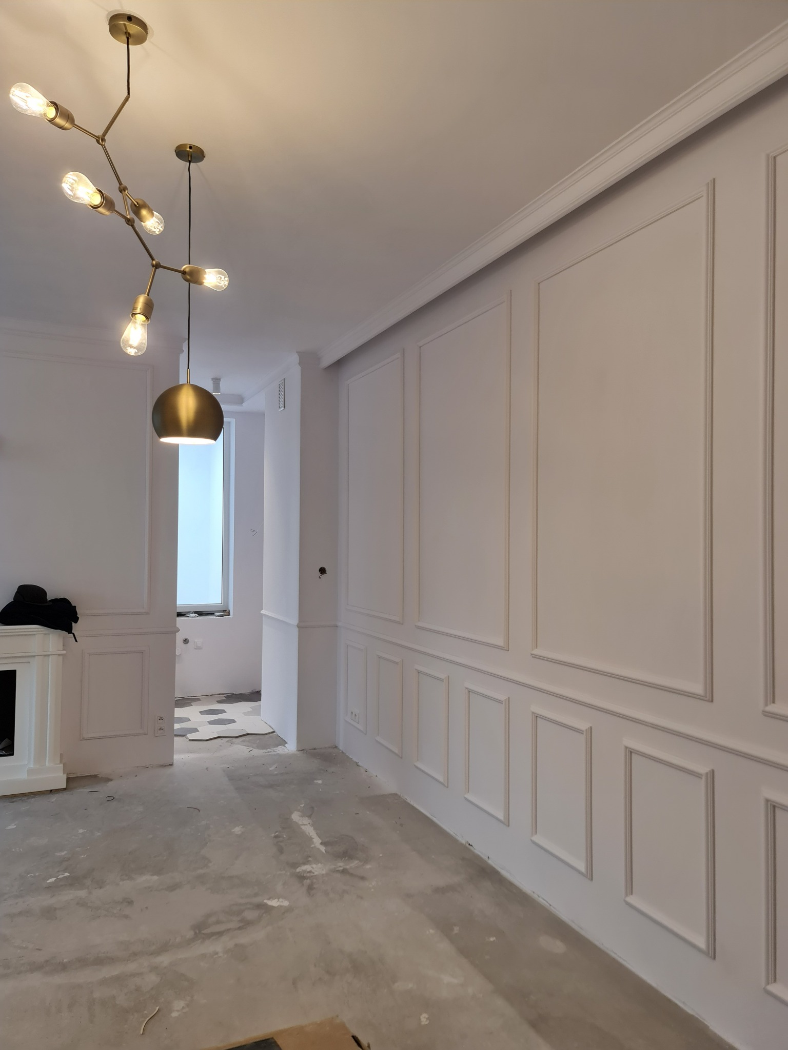 Pokój w trakcie wykańczania z dekoracyjnymi panelami ściennymi w stylu klasycznym, nowoczesna lampa wisząca z żarówkami Edison, surowa betonowa podłoga, widoczne ślady prac wykończeniowych, w tle...