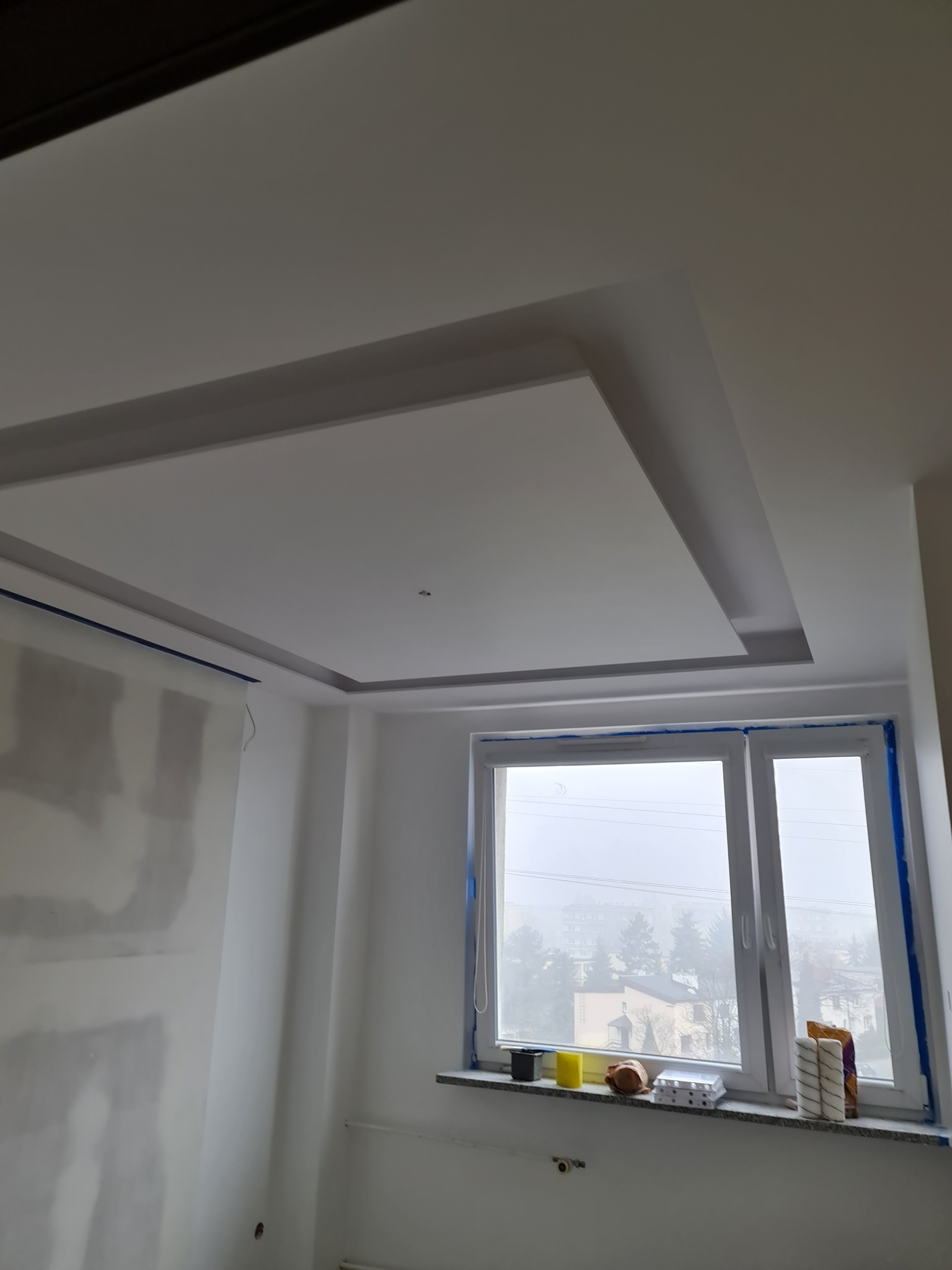 Wnętrze pokoju w trakcie remontu, widoczny podwieszany sufit z oświetleniem, ściany częściowo zagruntowane, zabezpieczone okno folią malarską, na parapecie wałki malarskie i pojemniki z farbą.