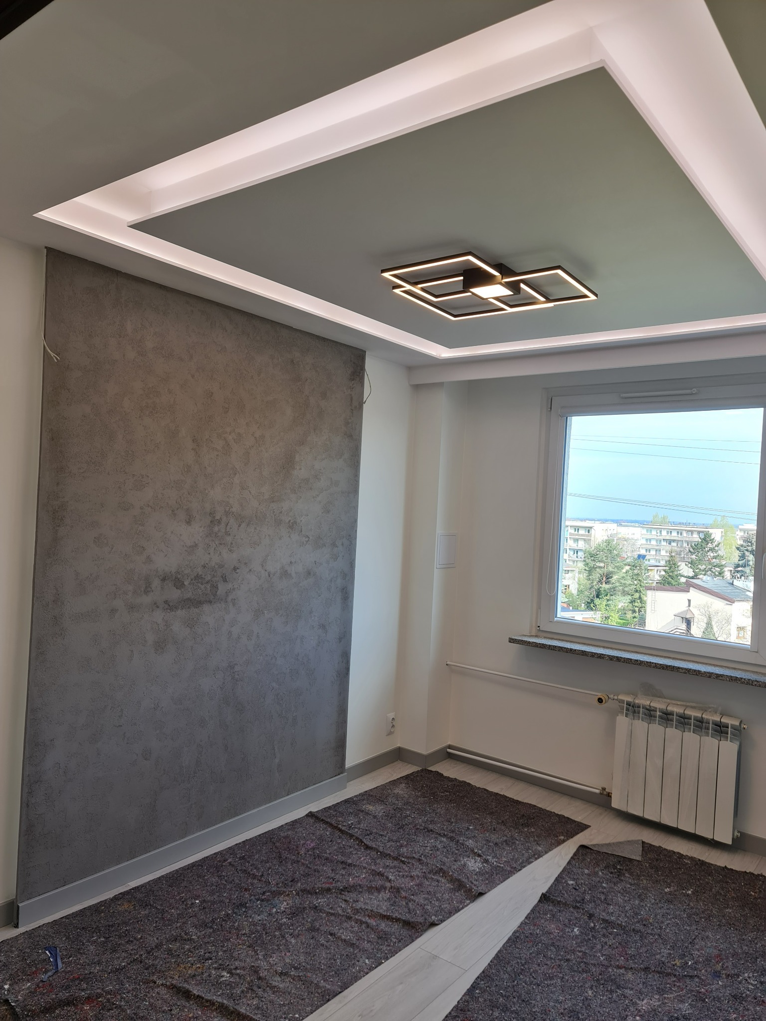 Pokój po remoncie: ściana z efektem betonu, nowoczesna lampa sufitowa z podświetleniem LED, okno z widokiem na miasto, zabezpieczona podłoga.