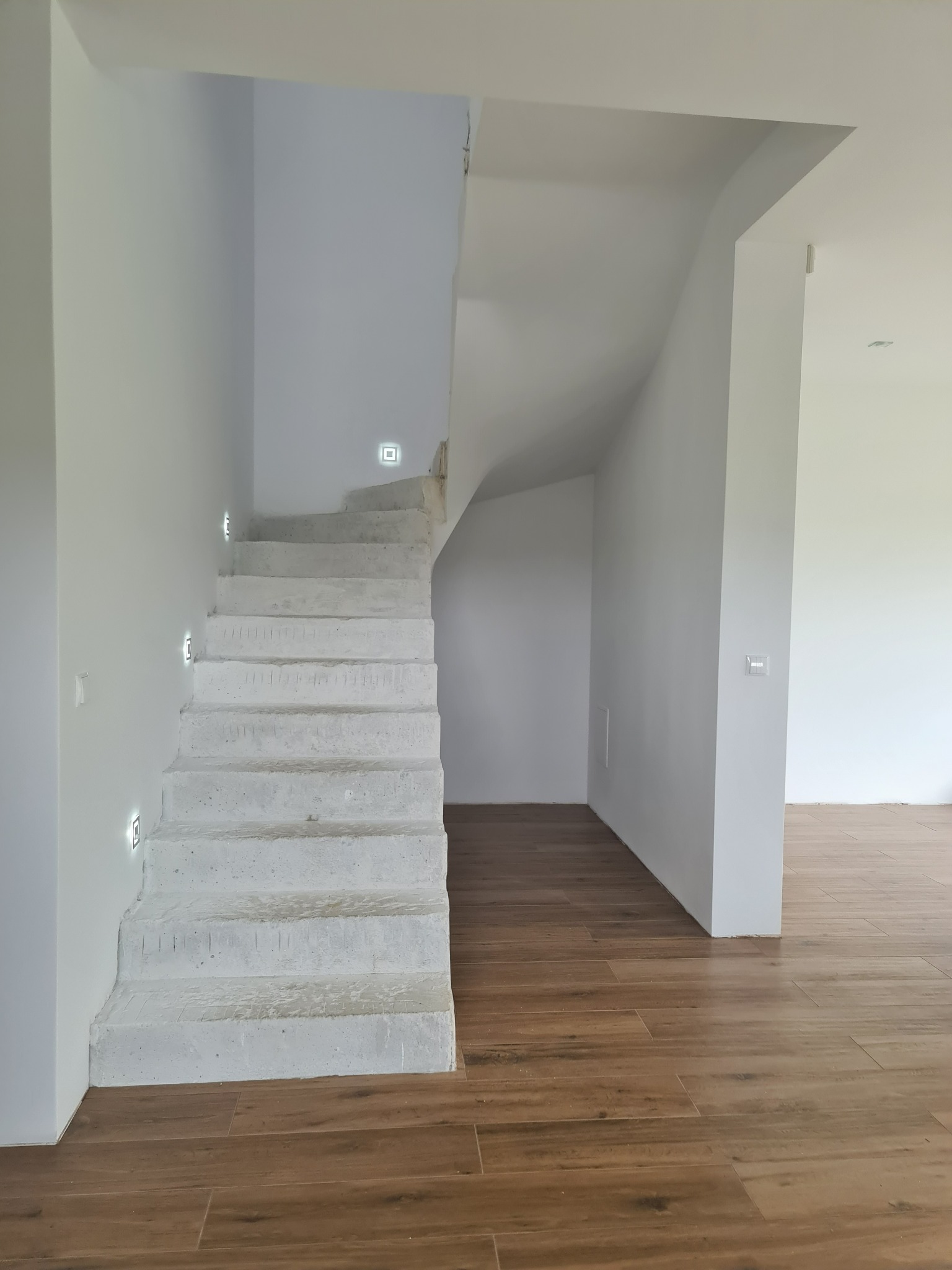 Surowe betonowe schody z wbudowanym oświetleniem LED na białej ścianie, brązowa podłoga imitująca drewno, widok na jasne wnętrze domu.