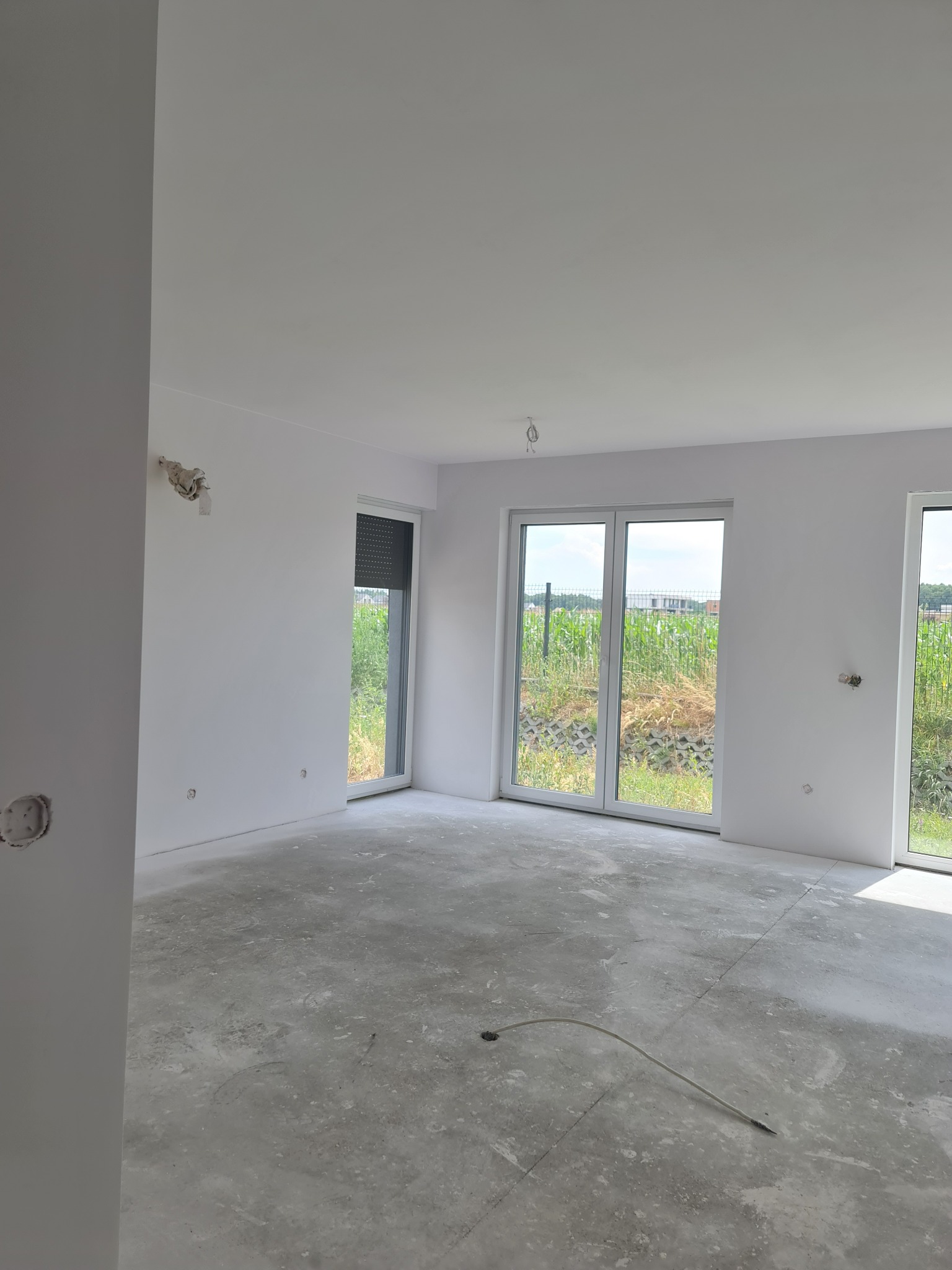 Surowe wnętrze z białymi ścianami, betonową podłogą, dużymi oknami z widokiem na zewnątrz i wystającymi kablami ze ściany, w trakcie prac wykończeniowych.