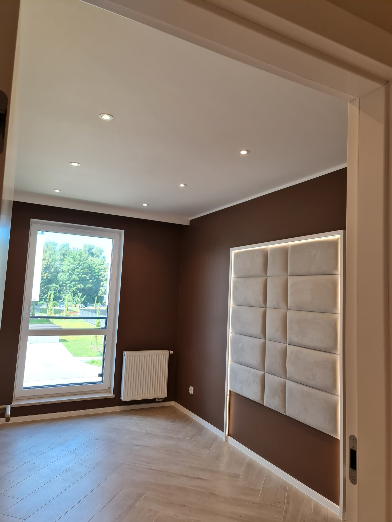 Pokój z brązowymi ścianami, panel tapicerowany z podświetleniem LED, parkiet w jodełkę, białe okno z widokiem na zieleń, grzejnik pod oknem, oświetlenie sufitowe punktowe.