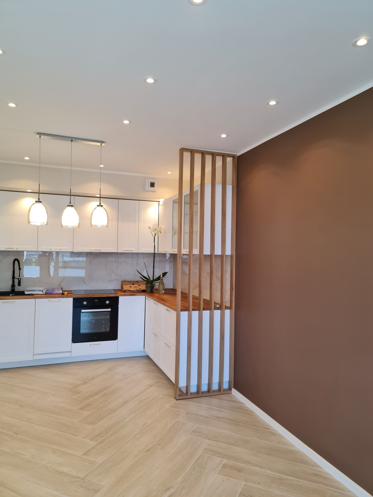 Nowoczesna kuchnia z białymi szafkami, drewnianym blatem, czarną armaturą i podłogą ułożoną w jodełkę, oddzielona od brązowej ściany ażurową przegrodą z jasnego drewna.