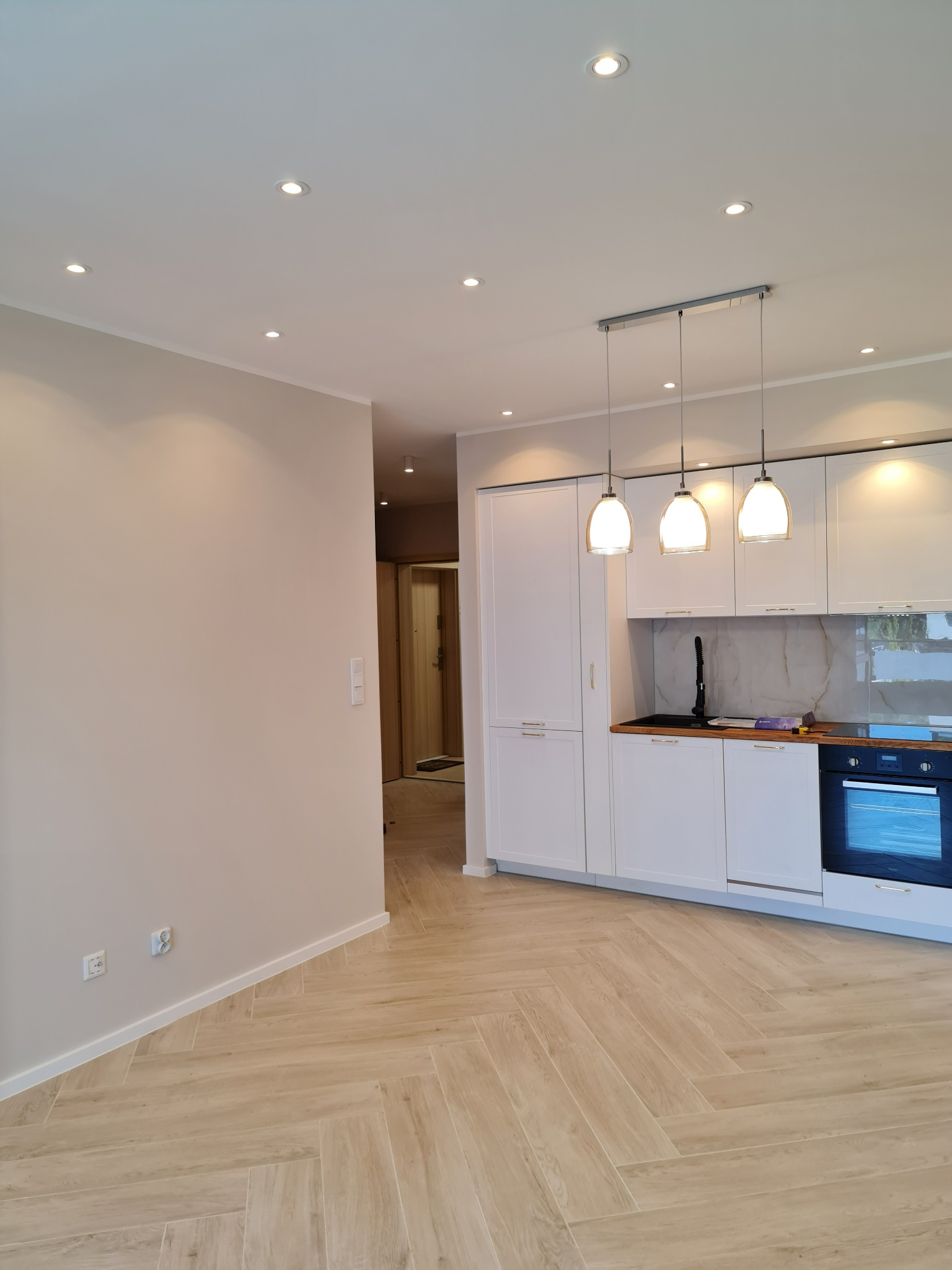 Jasna, nowocześnie wykończona kuchnia z białymi szafkami, drewnianym blatem i podłogą ułożoną w jodełkę, oświetlona wiszącymi lampami i halogenami.