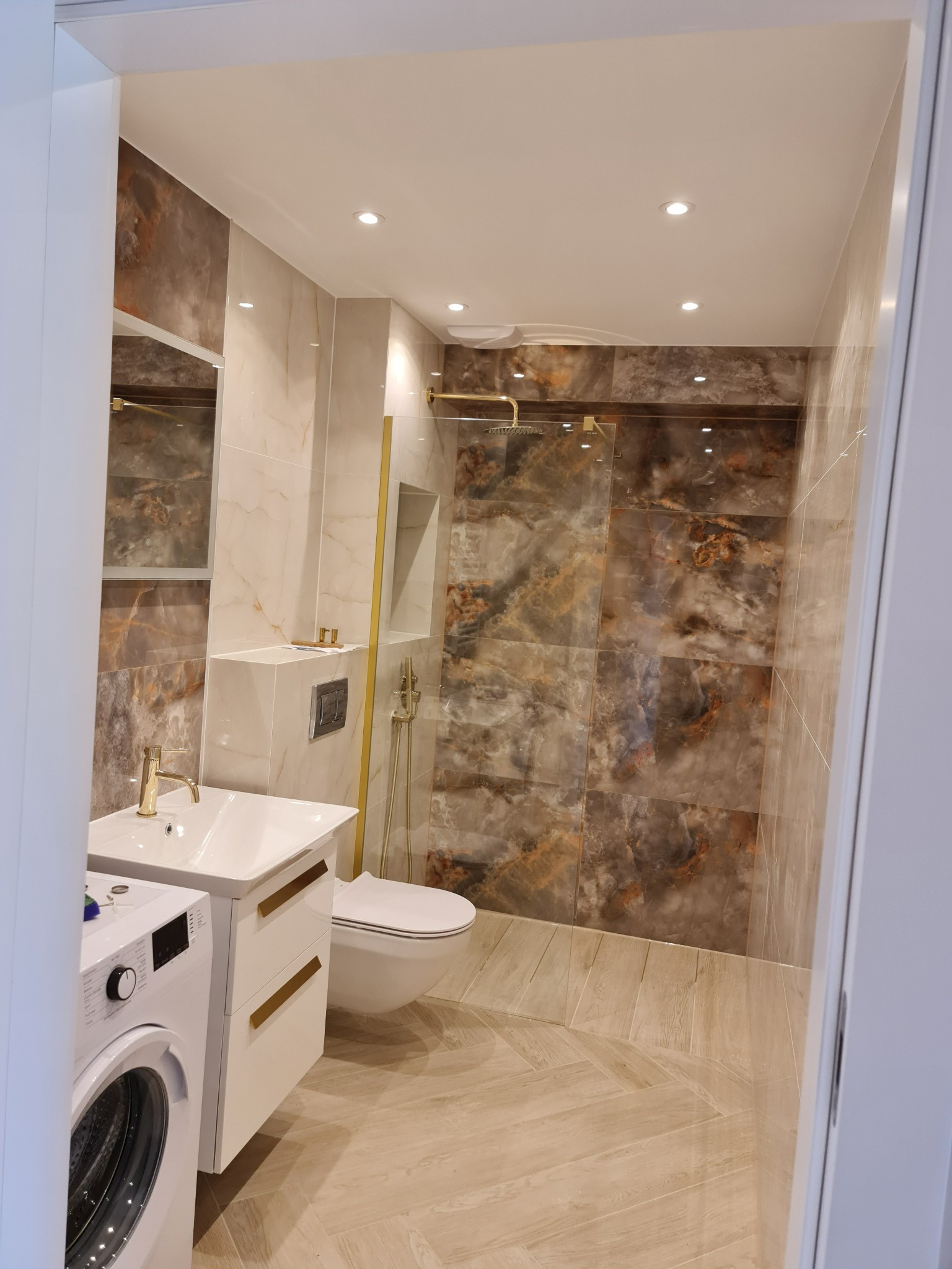 Nowoczesna łazienka z marmurowymi płytkami w odcieniach brązu i beżu, armaturą w kolorze złota, białą umywalką z szafką i pralką obok, oraz podłogą w jasną jodełkę.