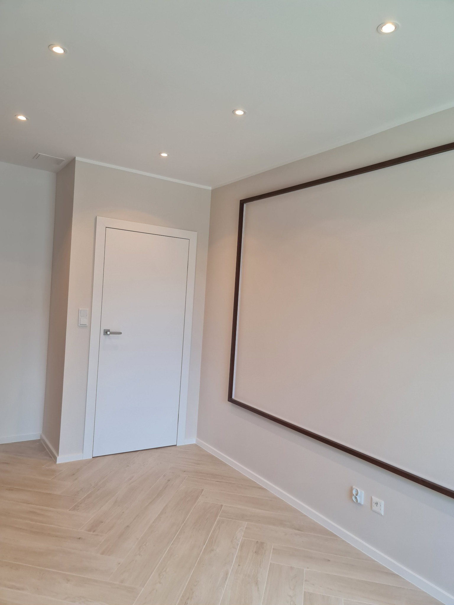 Jasne wnętrze po remoncie: parkiet ułożony w jodełkę, białe drzwi z srebrną klamką, ściana z ramą dekoracyjną, oświetlenie punktowe w suficie.