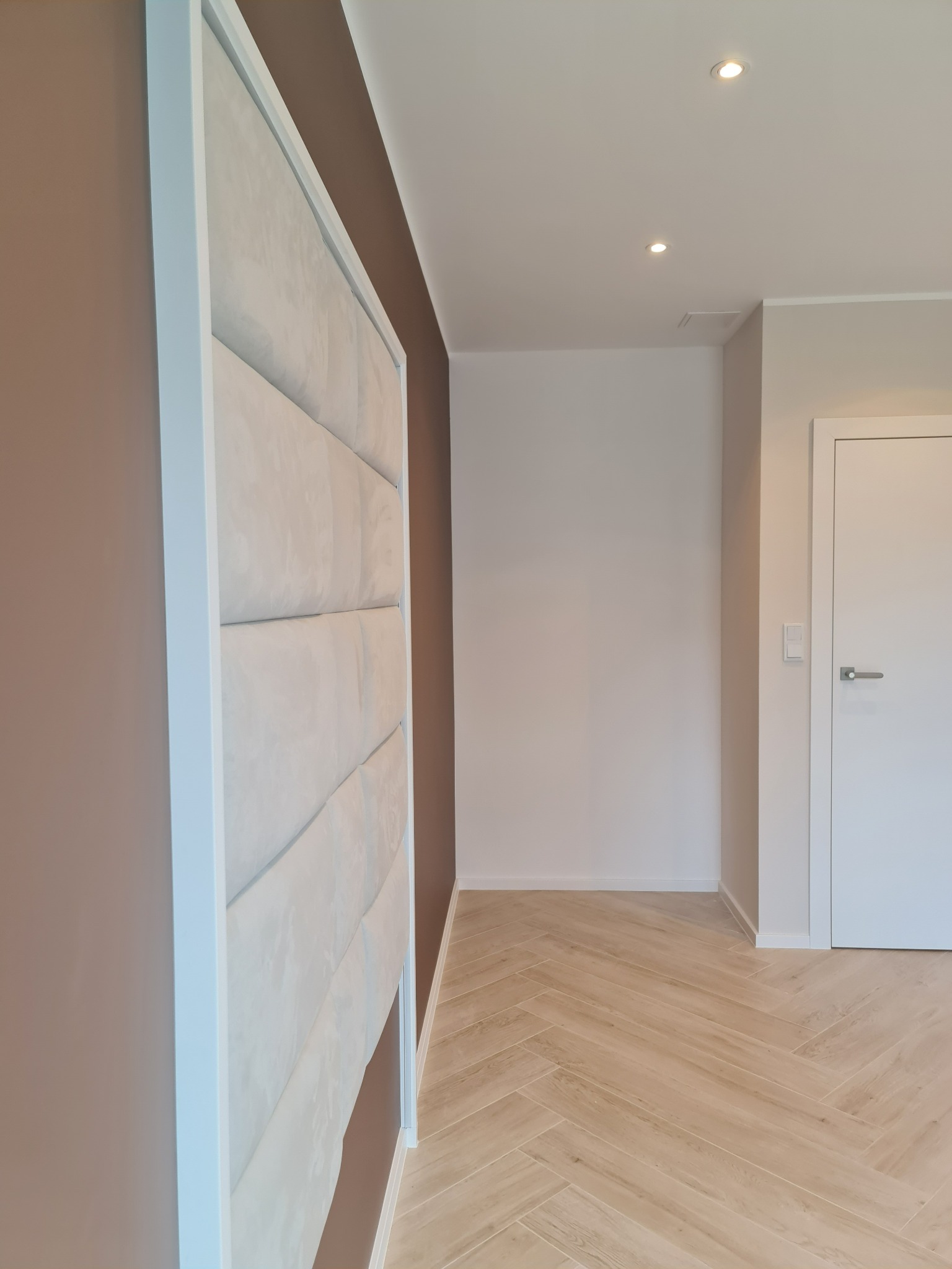 Beżowa tapicerowana ściana w białej ramie, brązowa ściana obok, parkiet w jodełkę, białe drzwi w tle, oświetlenie punktowe w suficie.