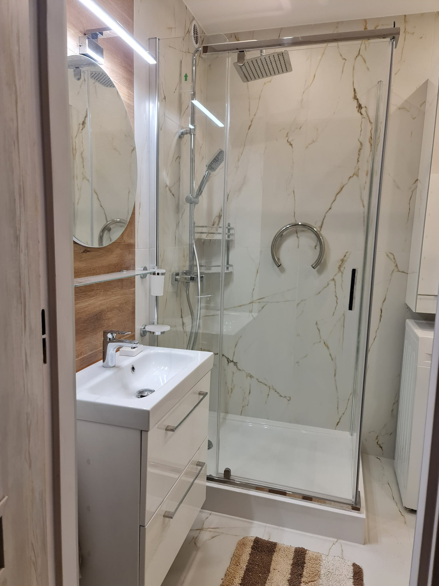 Nowoczesna łazienka z kabiną prysznicową typu walk-in, umywalką nablatową z szafką, okrągłym lustrem z podświetleniem LED i marmurowymi płytkami na ścianach.