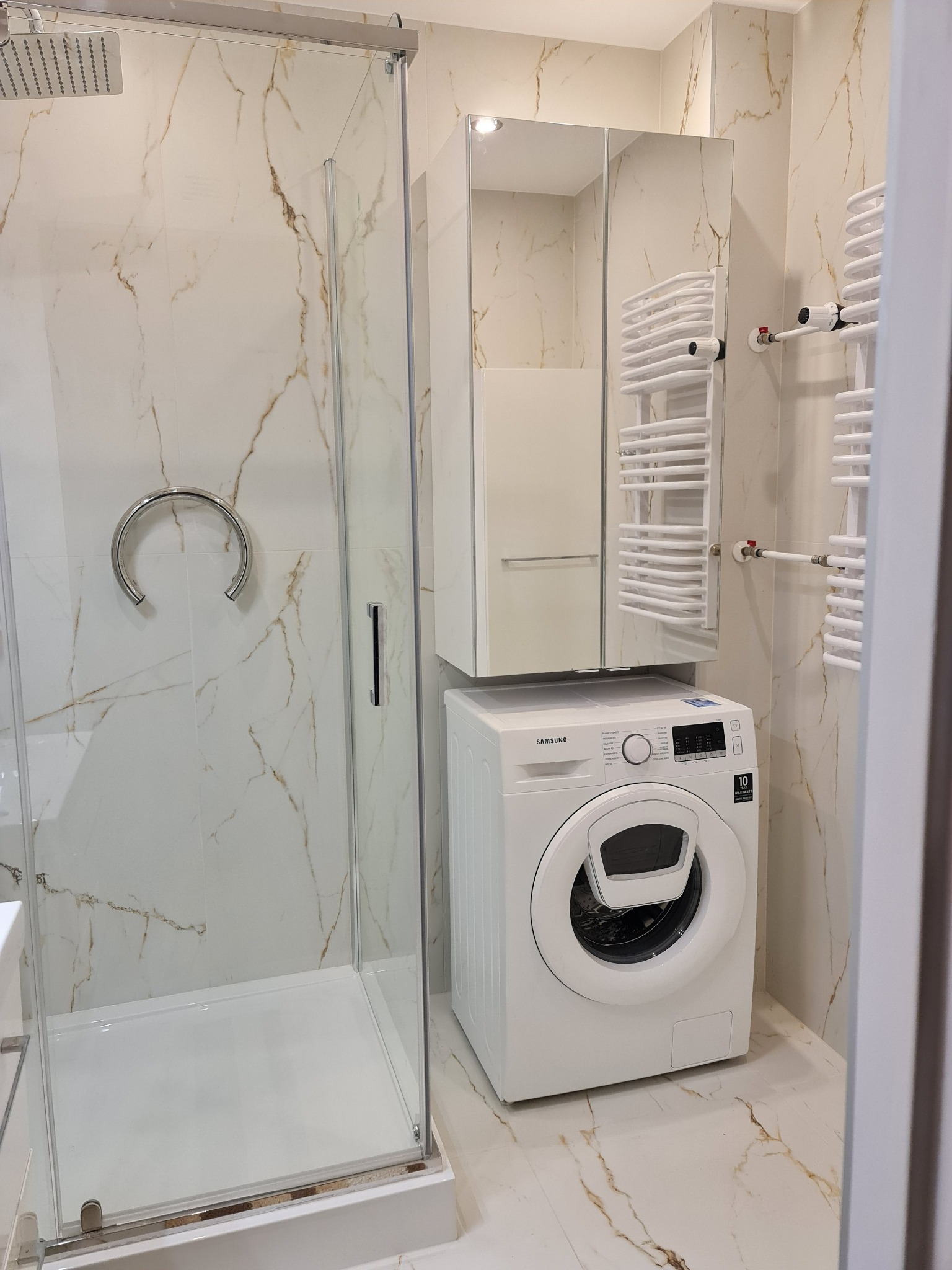 Nowoczesna łazienka z kabiną prysznicową, pralką Samsung i grzejnikiem drabinkowym na tle marmurowych płytek.