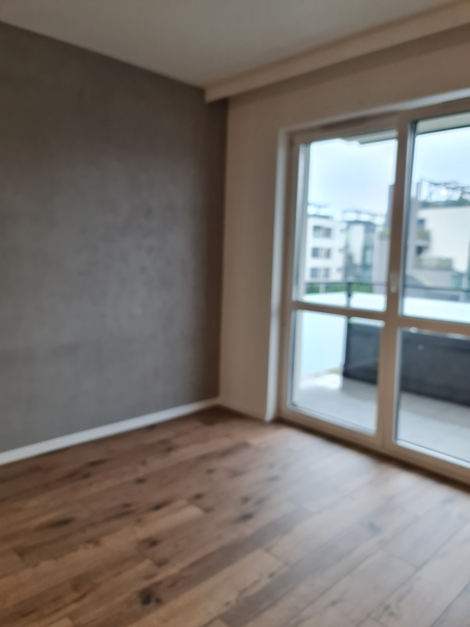 Pusty pokój z drewnianą podłogą i szarą ścianą, widok na balkon i budynki za oknem balkonowym, rozmyte zdjęcie.