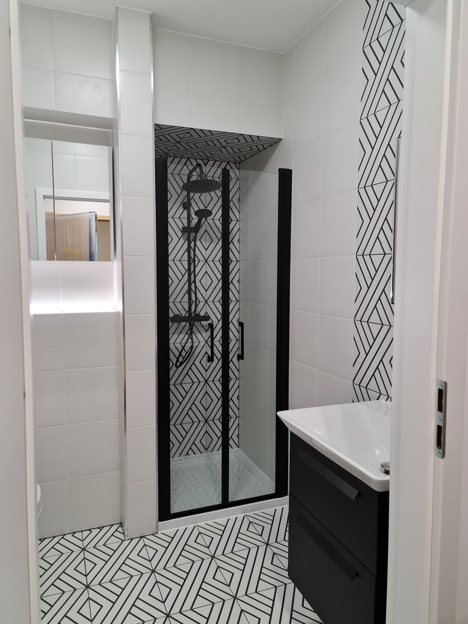 Nowoczesna łazienka z kabiną prysznicową w czarnej ramie, białe płytki z czarnym geometrycznym wzorem na podłodze, ścianie i suficie kabiny, czarna szafka pod umywalką.