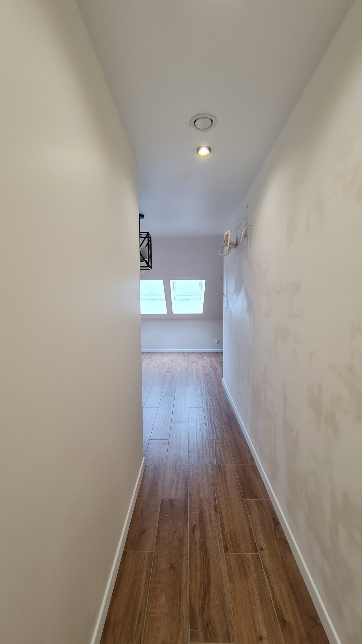 Wąski korytarz z brązową podłogą imitującą drewno, widoczne ślady po instalacji elektrycznej na ścianie, dwa okna dachowe w oddali i minimalistyczna lampa wisząca.