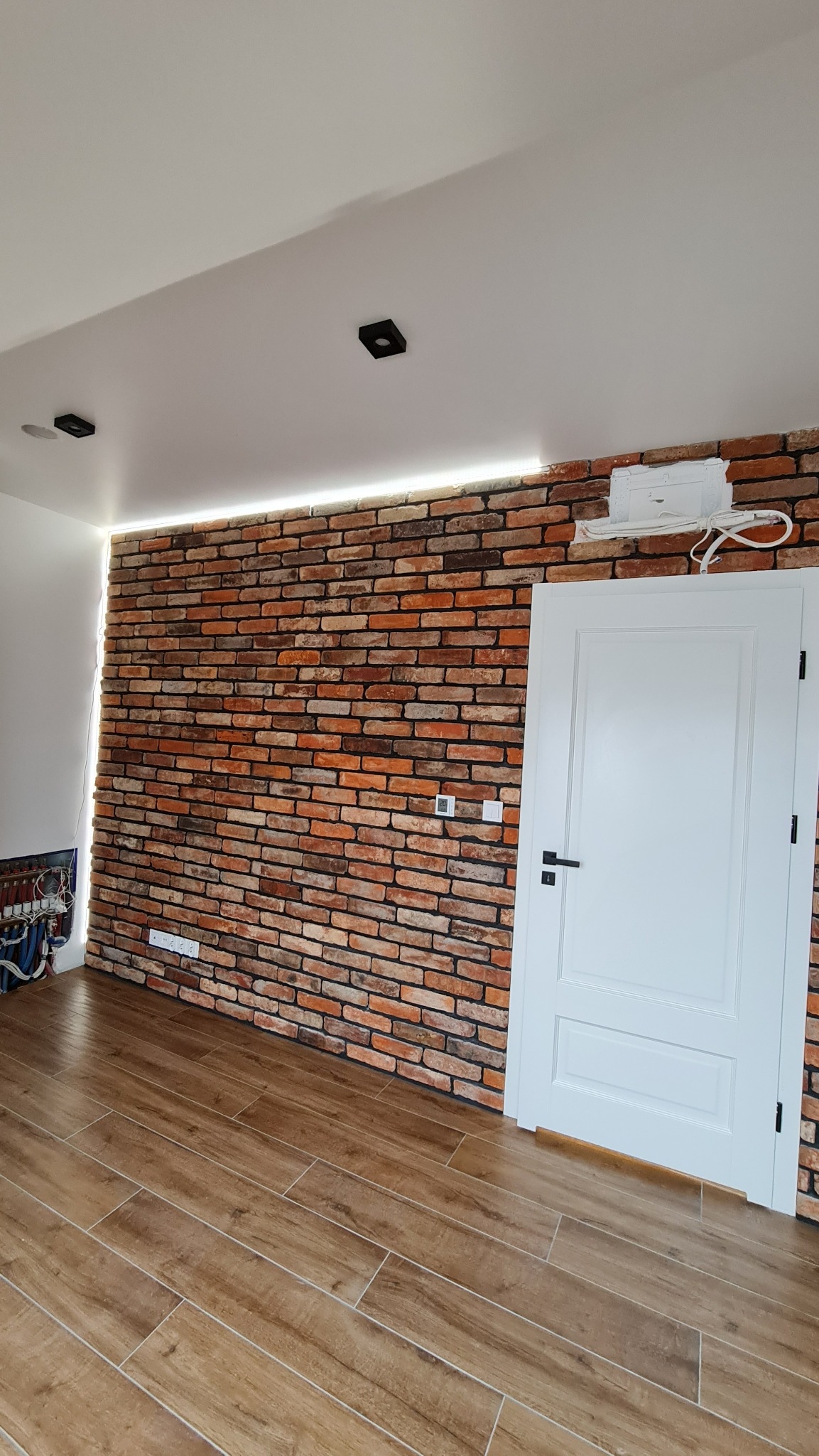 Ściana wykończona cegłą klinkierową z podświetleniem LED, białe drzwi z czarną klamką, podłoga z płytek imitujących drewno, widoczne elementy instalacji elektrycznej w rogu pomieszczenia.