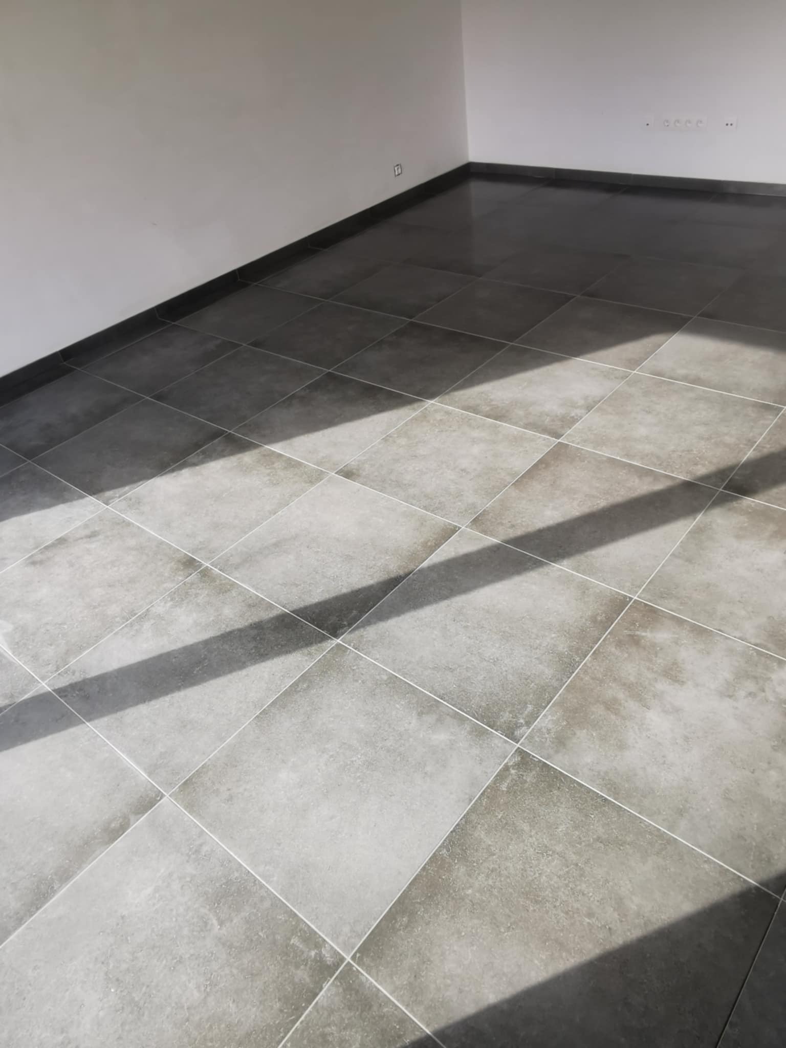 Ułożone szare płytki podłogowe w romby z widocznymi cieniami, jasne fugi, białe ściany w tle.