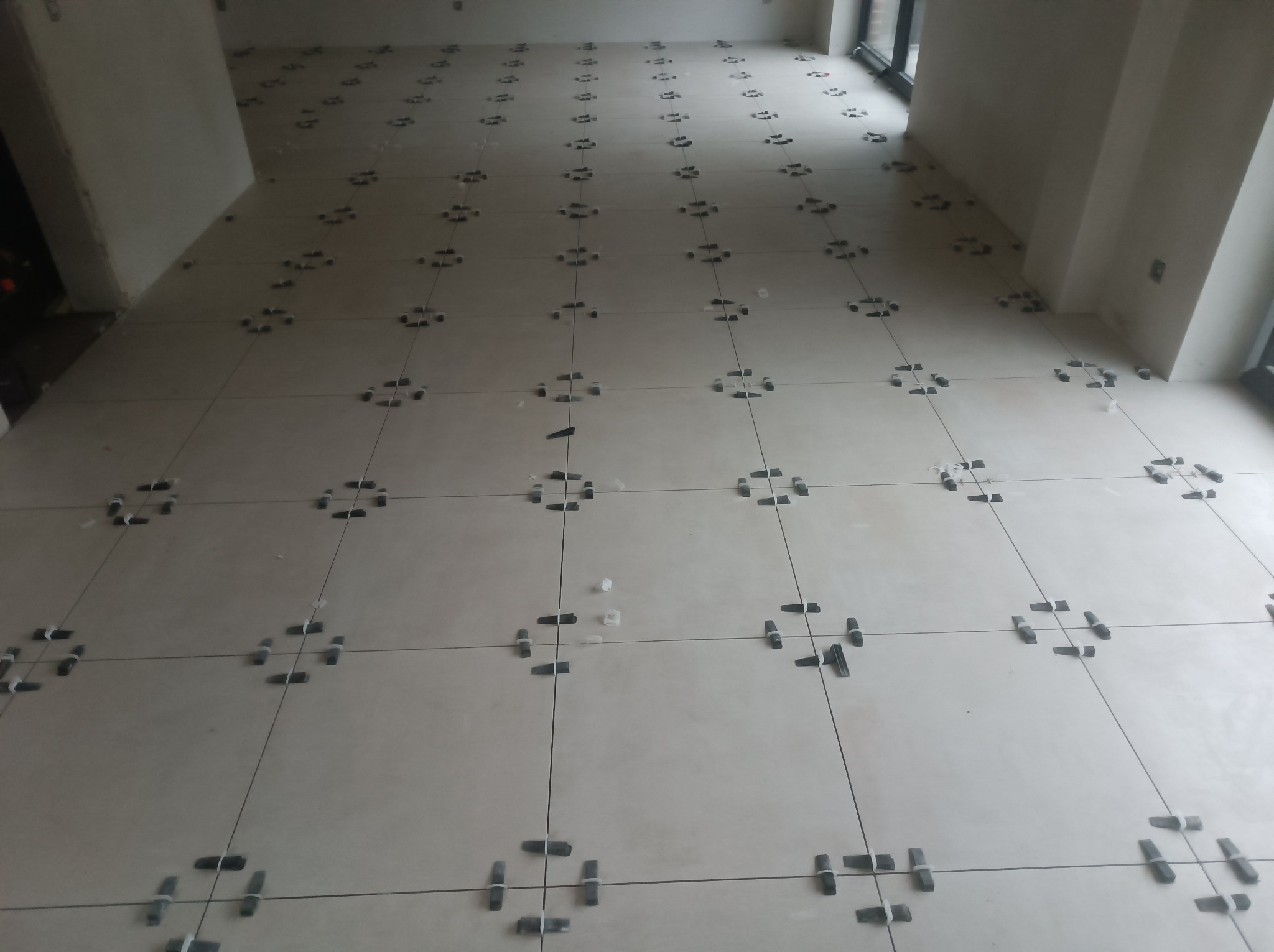 Jasne płytki podłogowe w trakcie układania w Obornikach, z użyciem czarnych i białych krzyżyków dystansowych, widoczne szare fugi i surowe ściany w tle.