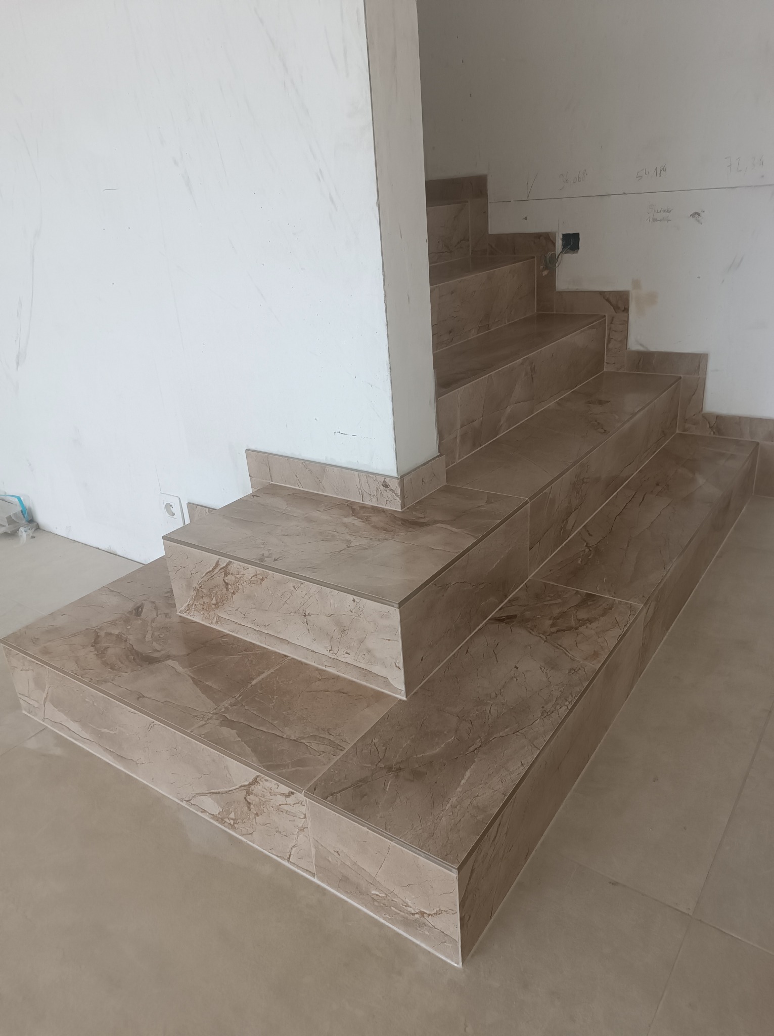 Wykończone schody wewnętrzne z płytek imitujących marmur, z widocznymi niedoskonałościami na ścianie i podłodze w trakcie remontu.