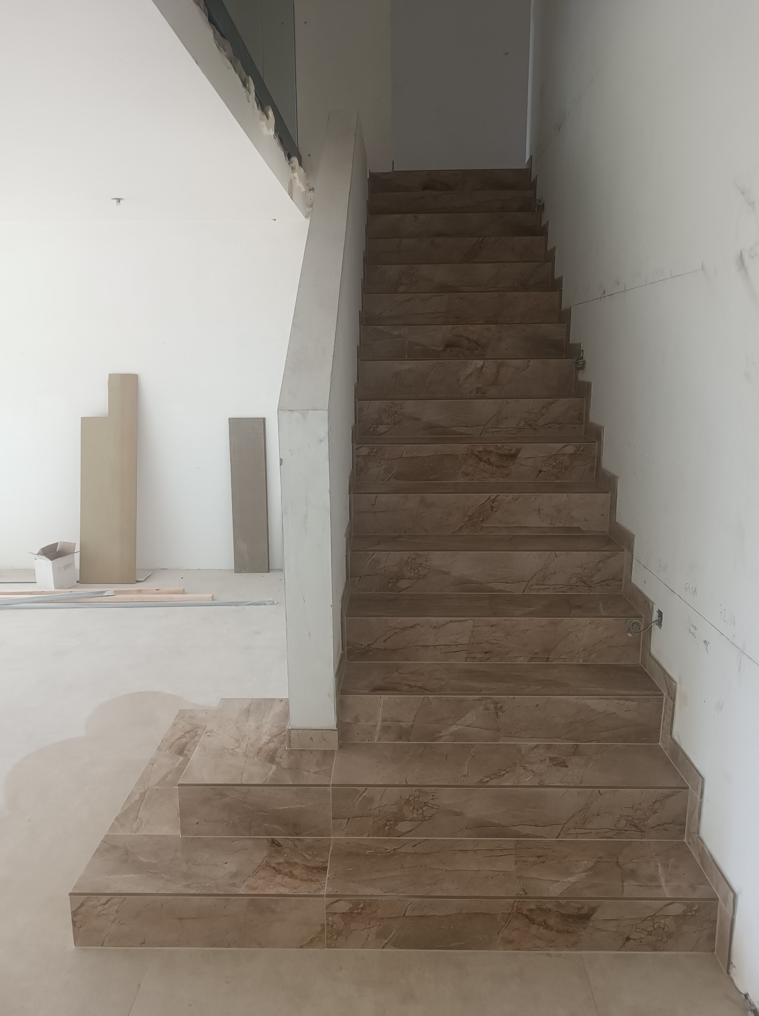 Schody wyłożone brązowymi płytkami imitującymi marmur, widoczne przygotowane płyty i karton na podłodze w surowym wnętrzu.