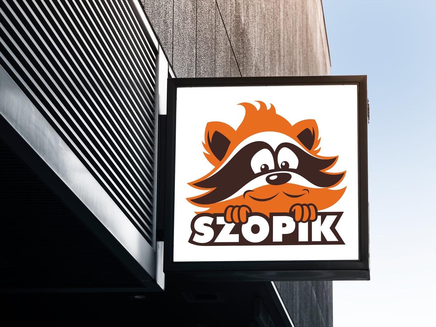 Kwadratowy szyld z podświetlanym logo przedstawiającym uśmiechniętego szopa w pomarańczowo-brązowych barwach, trzymającego napis 'SZOPiK', zamontowany na budynku z elementami w czarno-białe pasy.