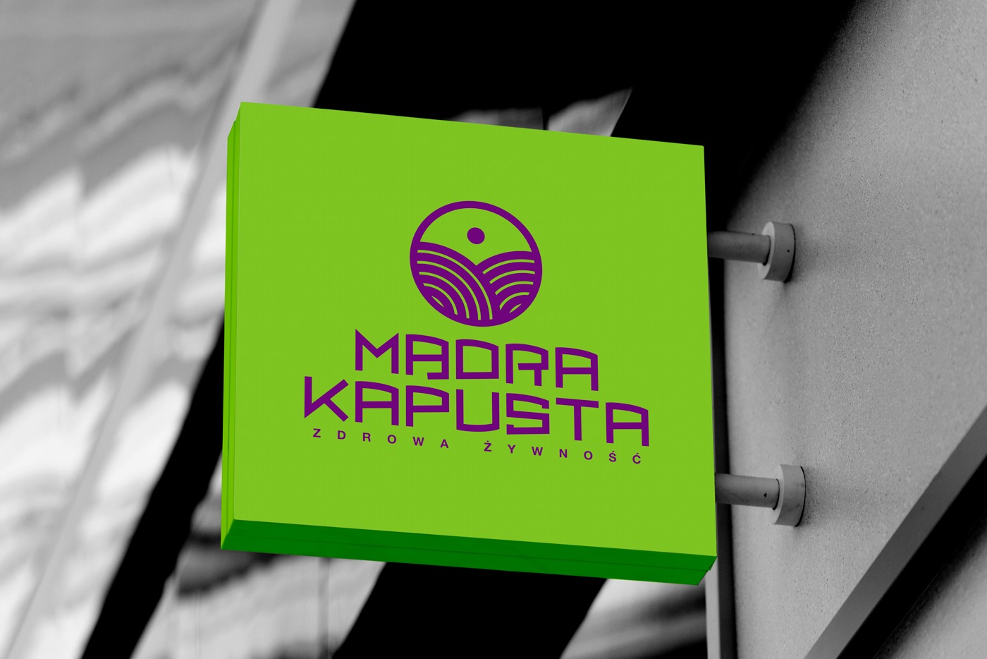 Zewnętrzny szyld reklamowy w kształcie kwadratu, z zielonym tłem i fioletowym logo oraz napisem 'Mądra Kapusta - zdrowa żywność', zamocowany do ściany budynku.