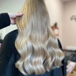 Długie, blond włosy z delikatnymi falami, widok z tyłu, dłoń stylisty poprawia fryzurę, czarny fartuch ochronny.