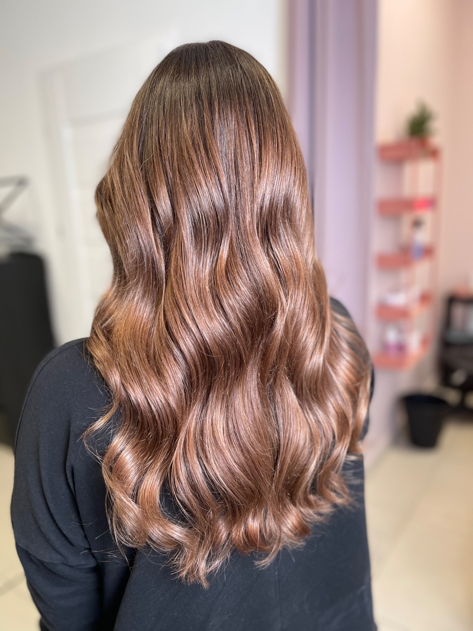 Długie, falowane włosy w odcieniach brązu z subtelnymi refleksami, widoczne od tyłu, na tle jasnego wnętrza salonu z elementami dekoracyjnymi na półce.