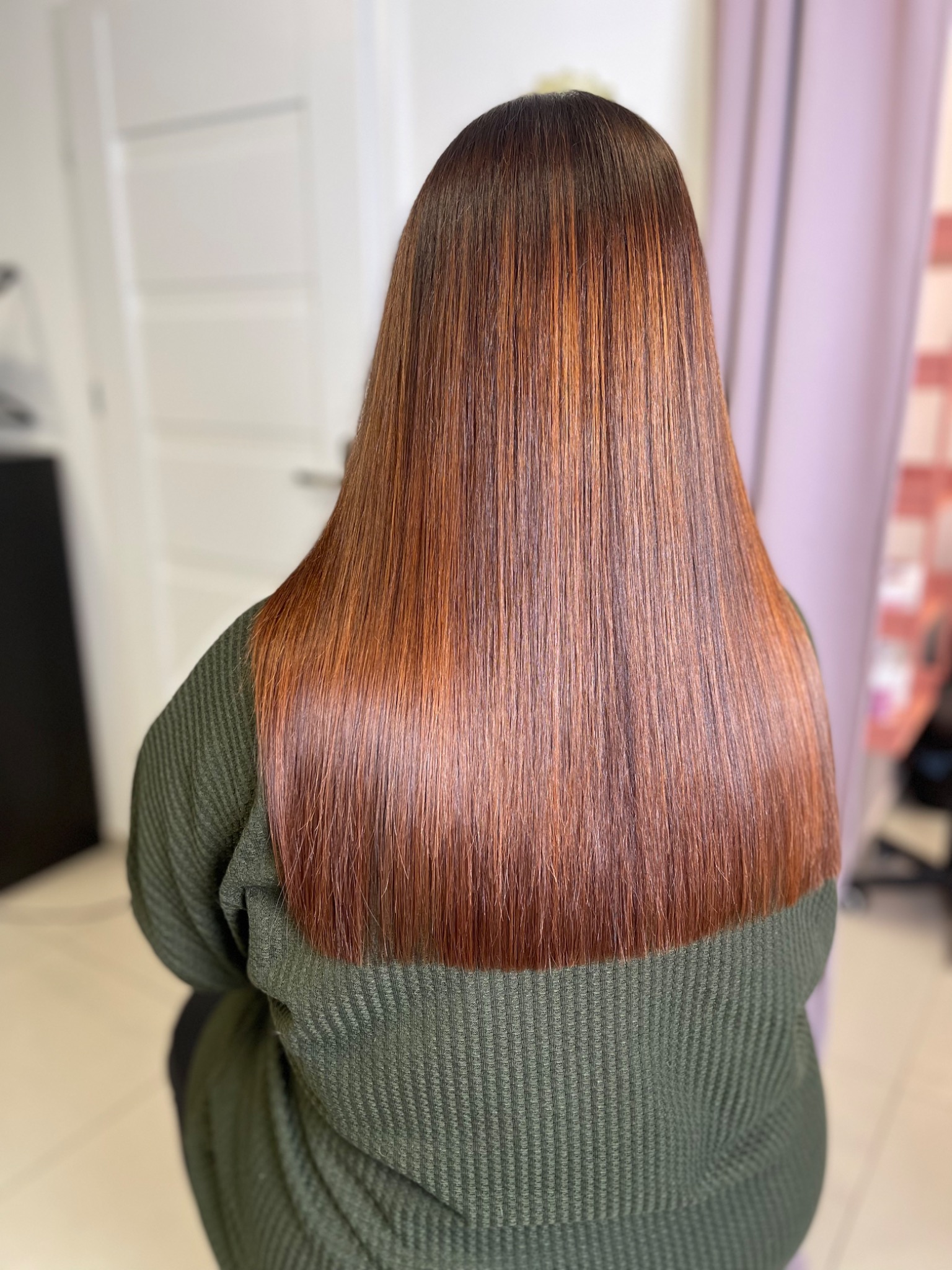 Długie, proste, brązowe włosy z refleksami, widoczne od tyłu, na tle jasnych drzwi i zasłony.