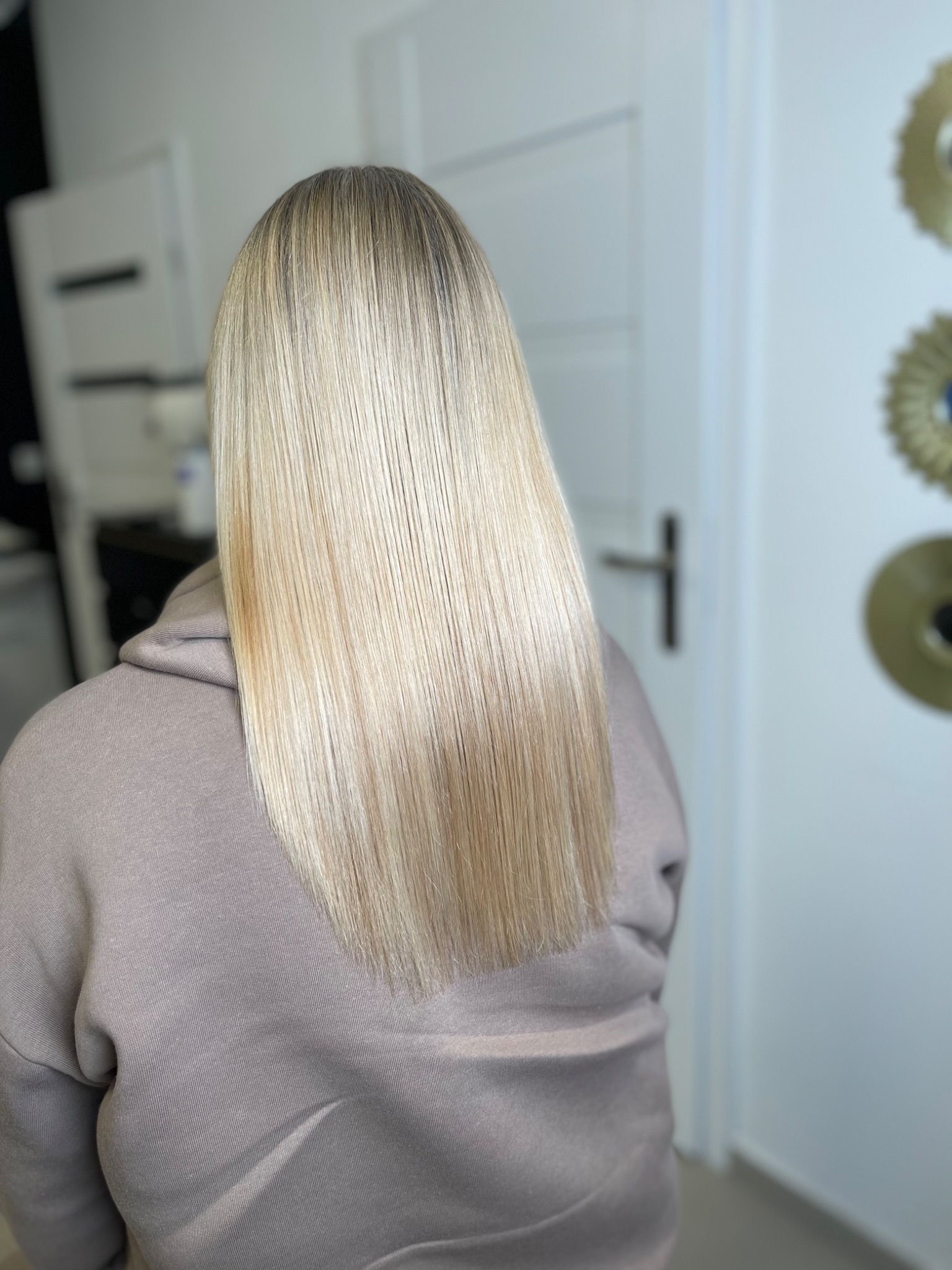 Proste, platynowe blond włosy do ramion, widok z tyłu, na tle białej ściany z dekoracjami.