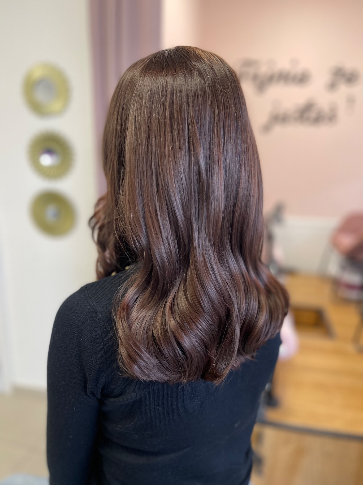 Błyszczące, brązowe włosy z delikatnymi falami, widok z tyłu, na tle jasnoróżowej ściany z napisem i dekoracyjnymi okrągłymi elementami.