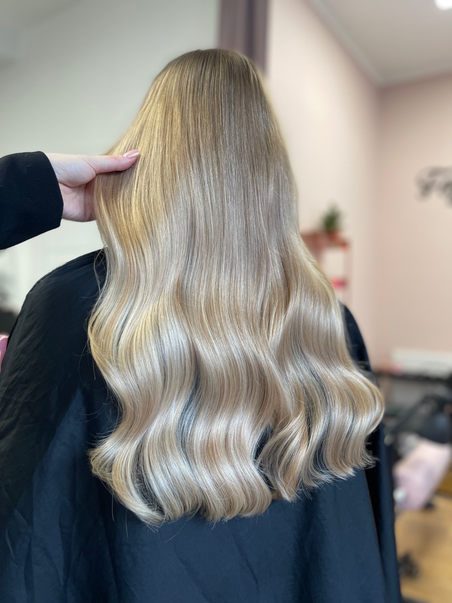 Długie, blond włosy z delikatnymi falami, widok z tyłu, dłoń stylisty poprawia fryzurę, czarny fartuch ochronny.