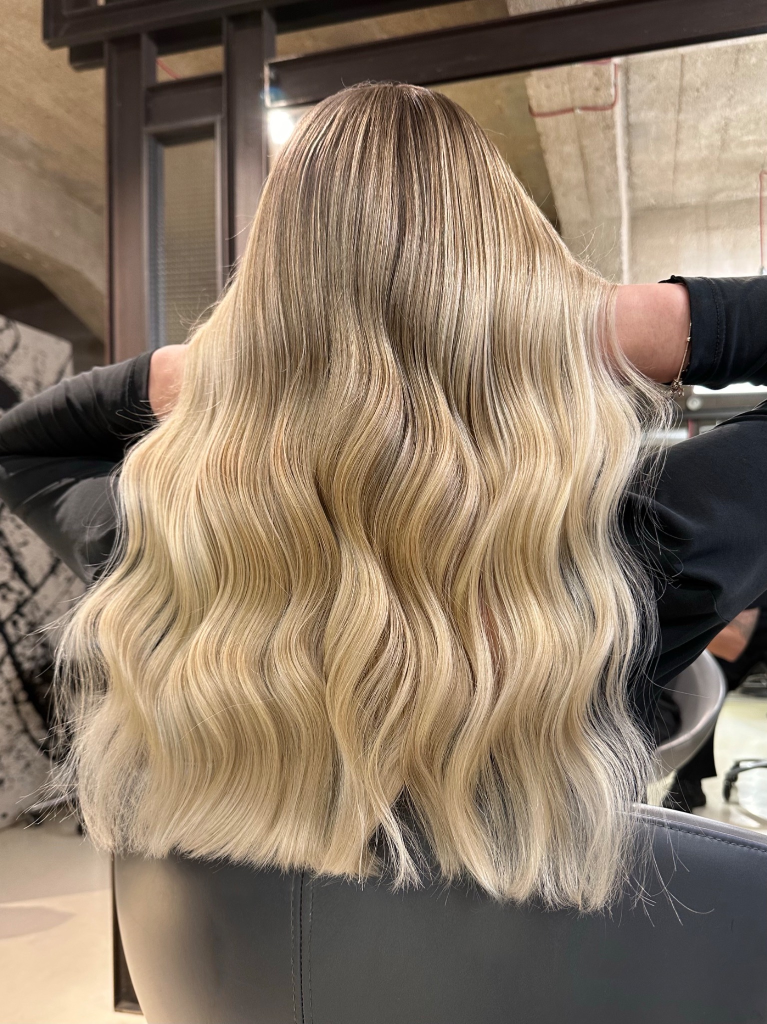 Długie, falowane blond włosy z refleksami, widok z tyłu, ręce uniesione za głową, w tle surowy betonowy sufit i metalowe elementy wystroju salonu fryzjerskiego.