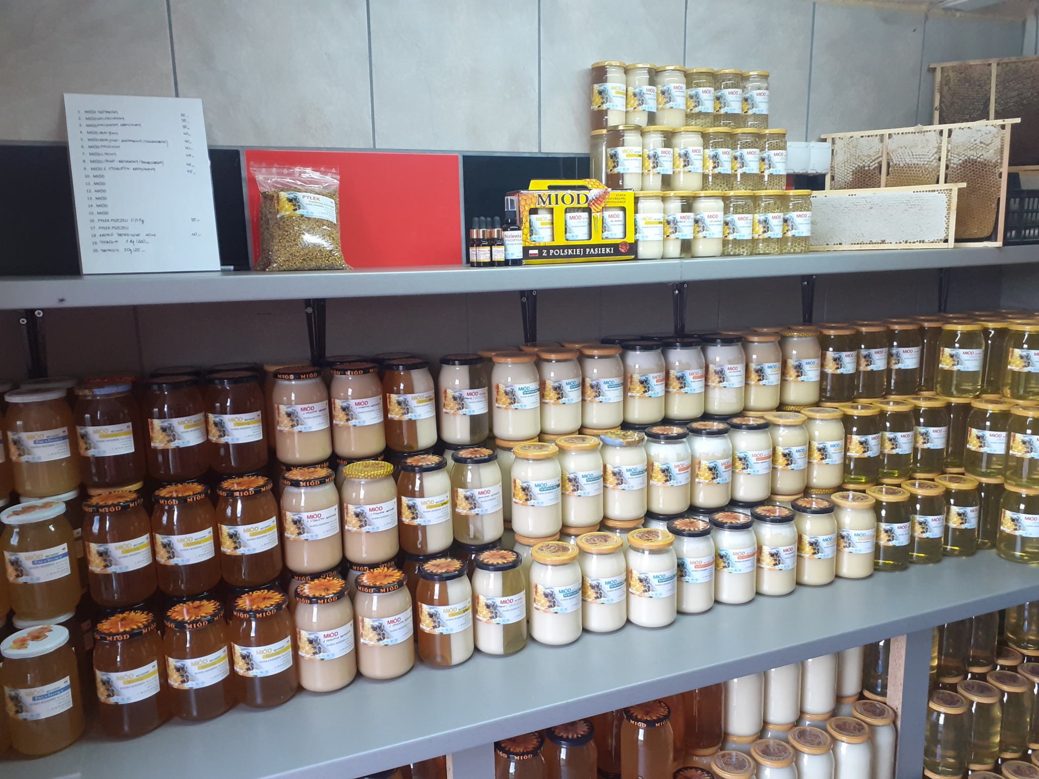 Półki sklepowe wypełnione słoikami z różnymi rodzajami miodu, pyłkiem pszczelim i zestawami prezentowymi, widoczna kartka z cenami.