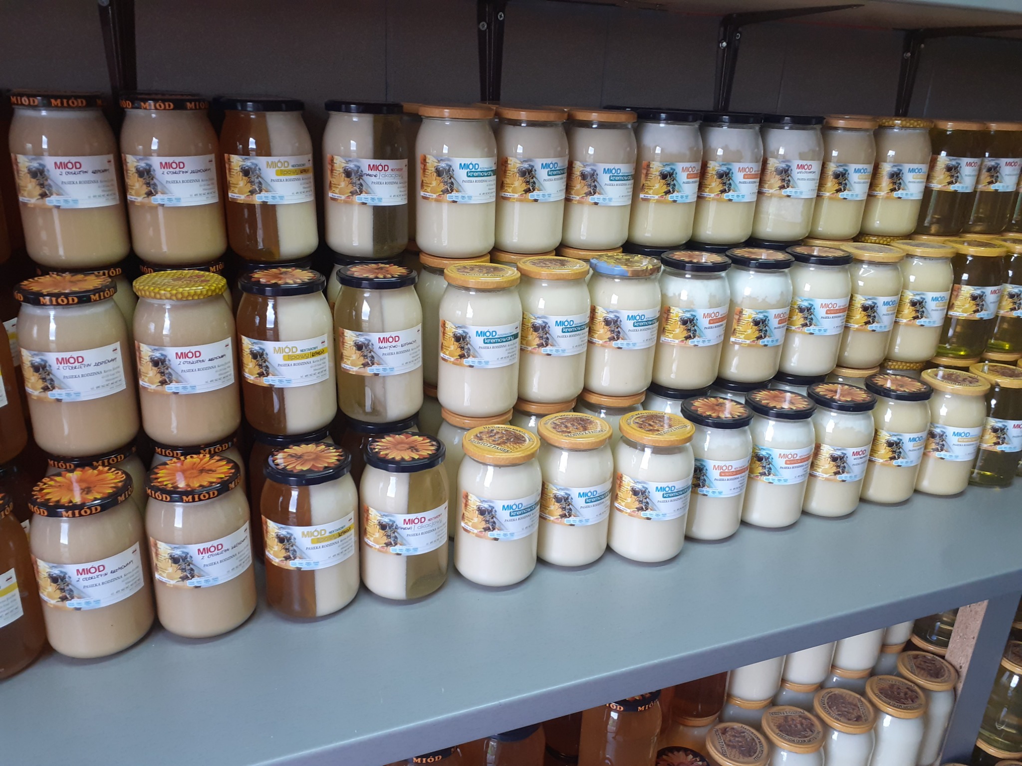 Półki w sklepie z różnymi rodzajami miodu w słoikach, etykiety z nazwą i grafiką pszczoły, nakrętki z motywem plastra miodu lub kwiatów.