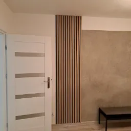 Nowoczesne wnętrze z drewnianymi lamelami na ścianie, betonem architektonicznym, białą, częściowo otwartą, drzwią z przeszkleniami i małym stolikiem.