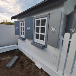 AGO Adam Grabowski - Biało-szara drewniana budowla z oknami i szarymi okiennicami, ozdobiona motywami zwierząt, otoczona ziemią i fragmentem białego płotu.