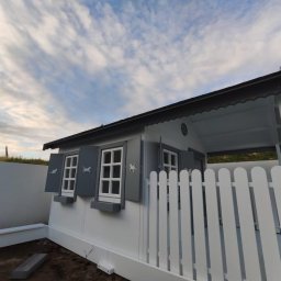 AGO Adam Grabowski - Biało-szara, drewniana altana dla dzieci z motywem zwierząt na okiennicach, otoczona białym płotem i murem, na tle pochmurnego nieba. Widok z dołu.