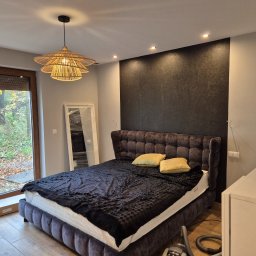 AGO Adam Grabowski - Sypialnia z łóżkiem z wezgłowiem, lampą z rattanu i akcentową ścianą w odcieniach szarości. Widoczne okno z widokiem na jesienny ogród i elementy dekoracyjne.