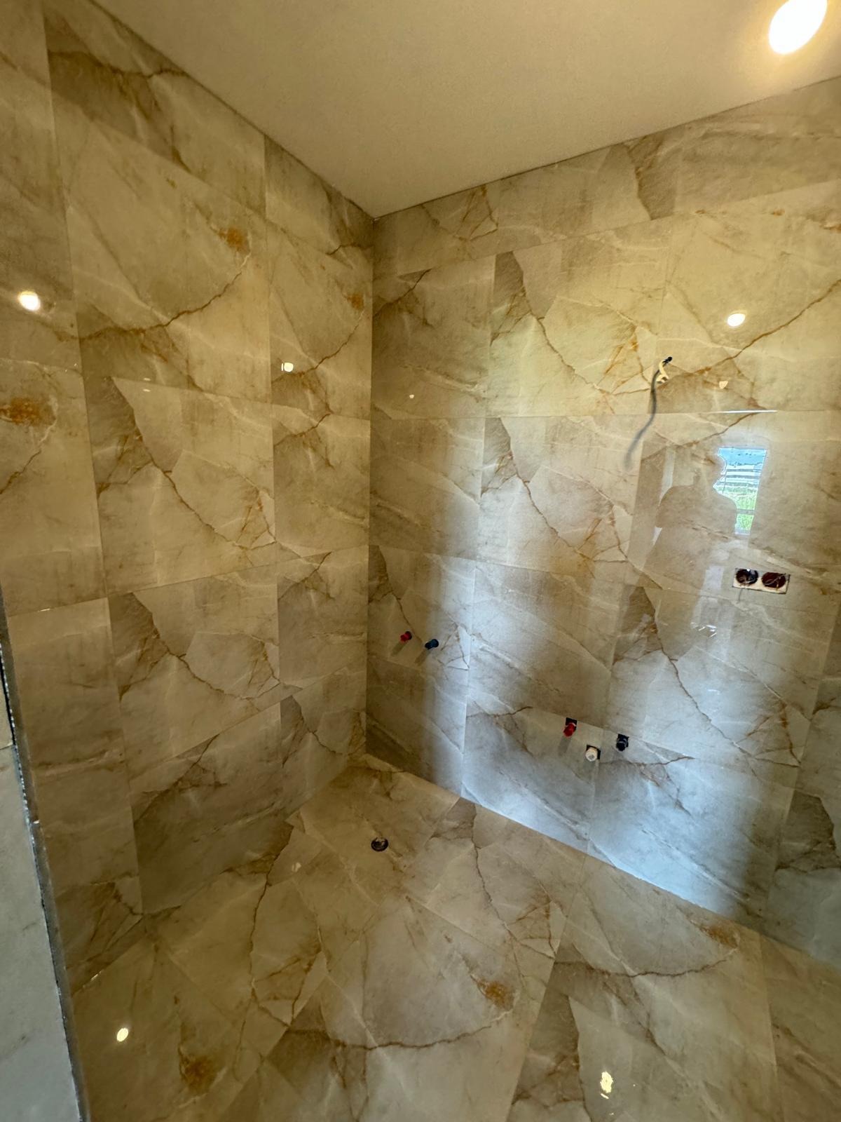 Wykończona kabina prysznicowa z beżowymi płytkami imitującymi marmur. Widoczne instalacje wodne i odpływ podłogowy. Nowoczesny design z połyskiem.