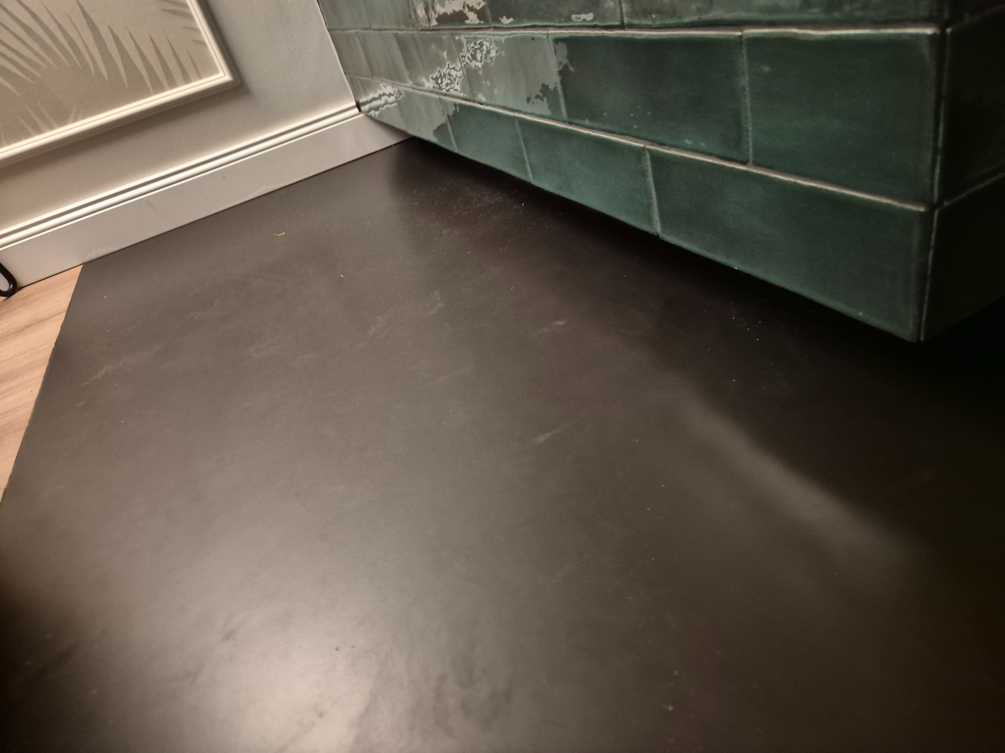 Ciemna, gładka posadzka z widoczną ścianą wyłożoną zielonymi, błyszczącymi płytkami. Fragment listwy przypodłogowej i ściany z dekoracyjnym wzorem w tle.