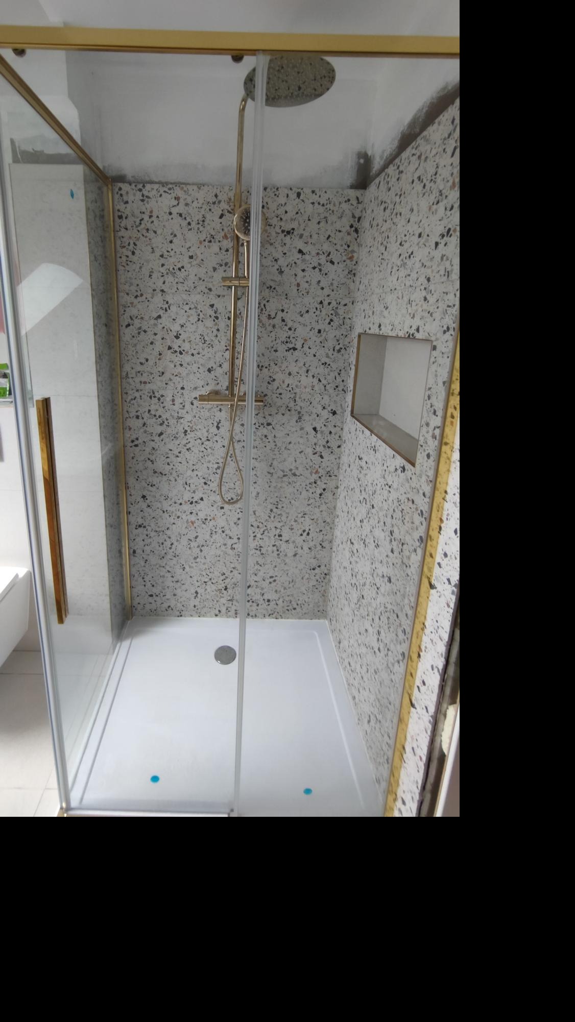 Kabina prysznicowa z brodzikiem i złotymi ramami, ściany wyłożone płytkami imitującymi lastryko, wnęka w ścianie. Nowoczesny design łazienki.