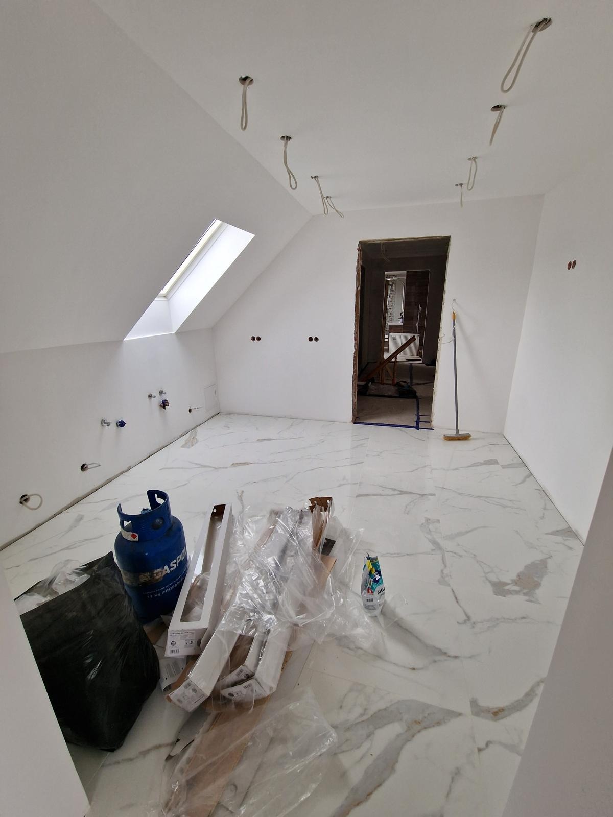Poddasze z oknem dachowym i marmurową posadzką w trakcie prac. Widoczne przygotowane materiały, butla gazowa i wystające kable. Stan surowy, przed wykończeniem.