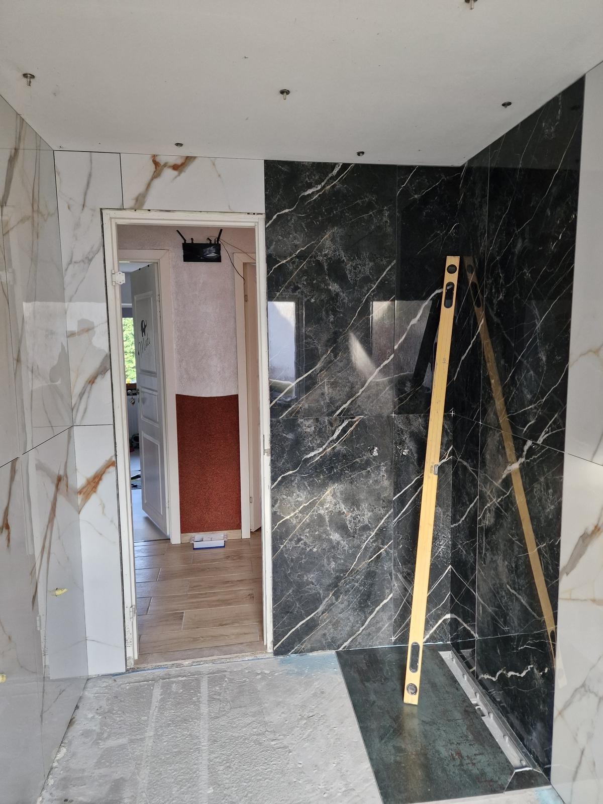 Częściowo wykończona łazienka z marmurowymi płytkami w odcieniach bieli i czerni. Poziomica oparta o ścianę. Widoczny odpływ liniowy. Otwór drzwiowy prowadzi do innego pomieszczenia.