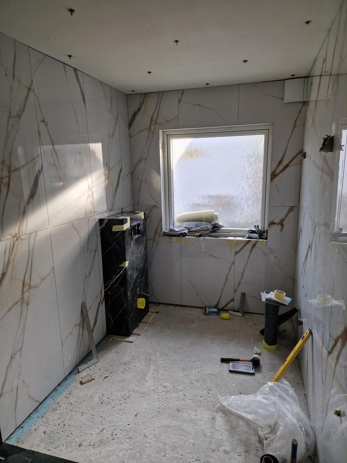 Wnętrze łazienki w trakcie prac wykończeniowych: marmurowe płytki na ścianach, zabudowa WC w czarnym marmurze, okno z widokiem na zewnątrz, narzędzia pracy na podłodze.