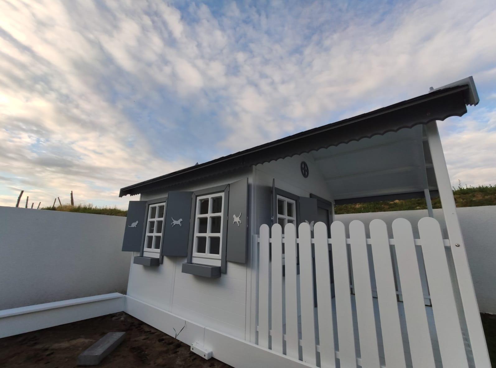 Biało-szara, drewniana altana dla dzieci z motywem zwierząt na okiennicach, otoczona białym płotem i murem, na tle pochmurnego nieba. Widok z dołu.