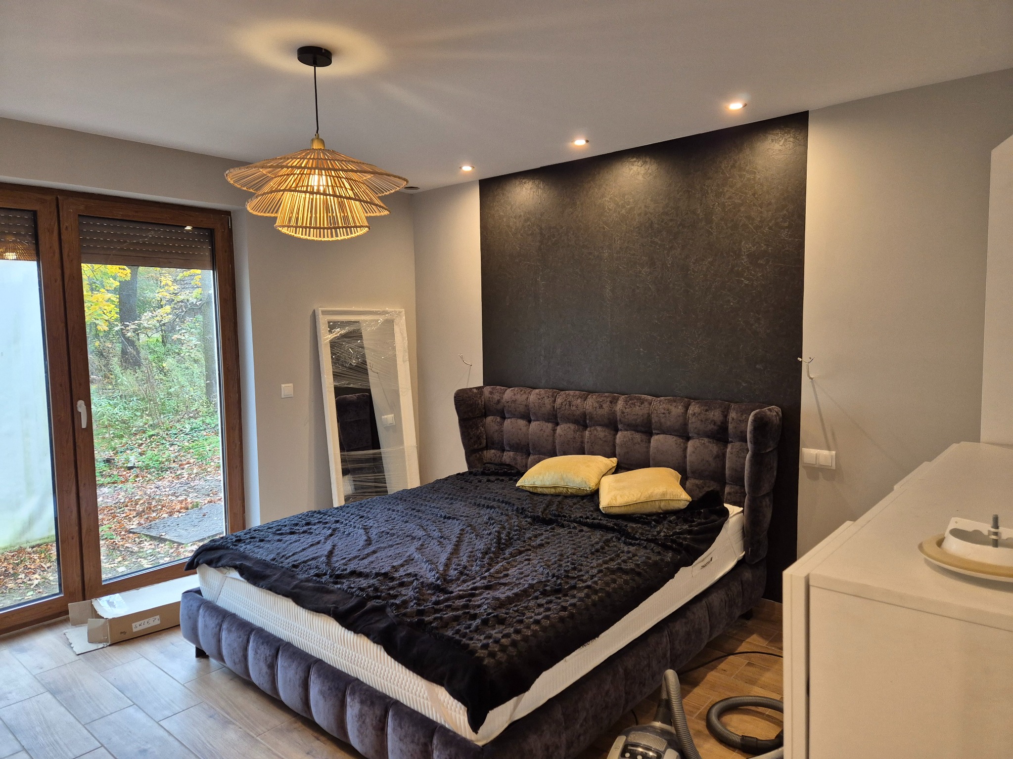 Sypialnia z łóżkiem z wezgłowiem, lampą z rattanu i akcentową ścianą w odcieniach szarości. Widoczne okno z widokiem na jesienny ogród i elementy dekoracyjne.