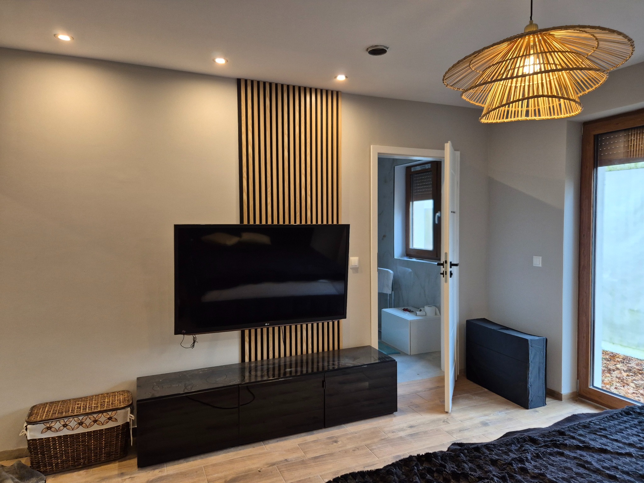 Nowoczesny salon z drewnianą ścianką działową za telewizorem, czarną szafką RTV i wiklinowym koszem. Widok na łazienkę przez otwarte drzwi.