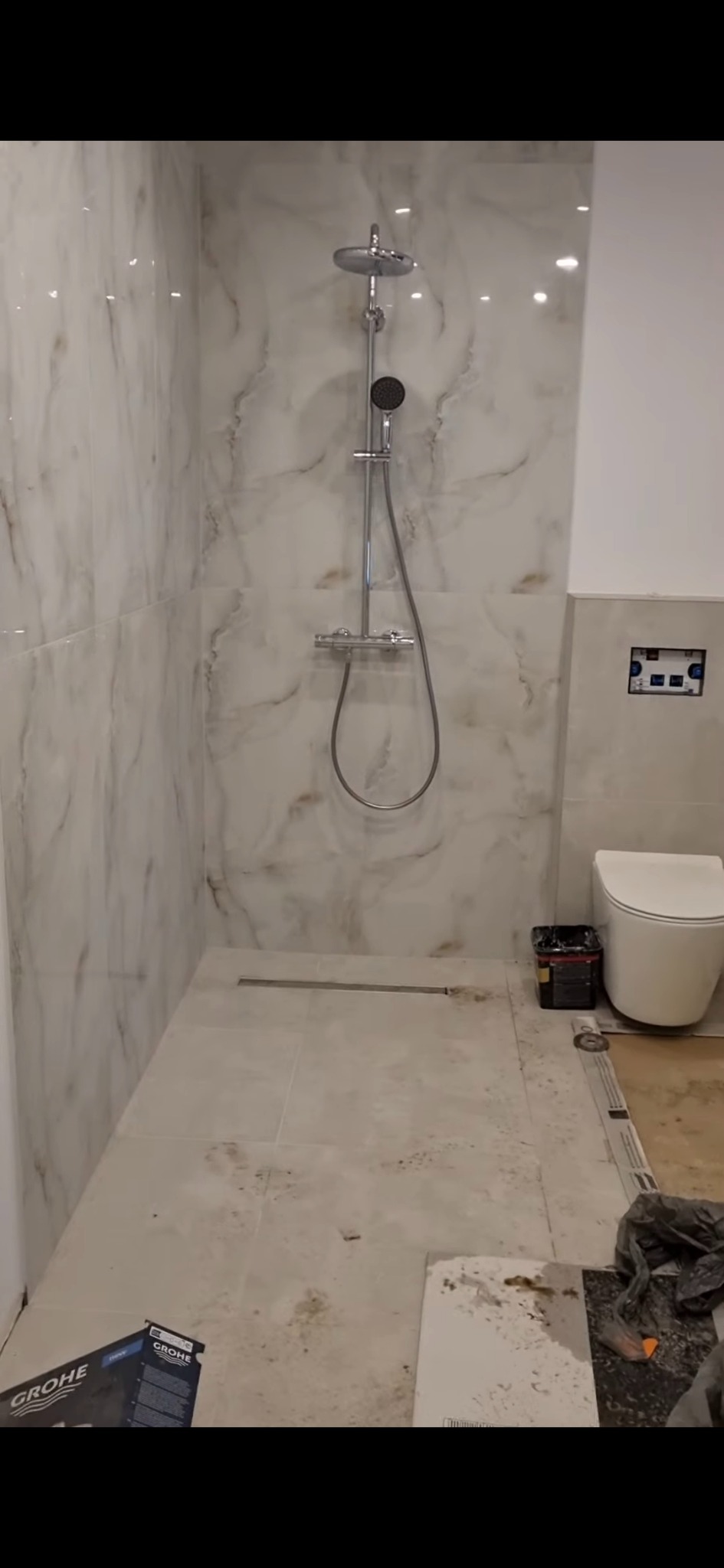 Łazienka w trakcie wykańczania: marmurowe płytki na ścianach, zamontowany prysznic, toaleta i widoczne narzędzia glazurnicze. Częściowo ułożona podłoga.