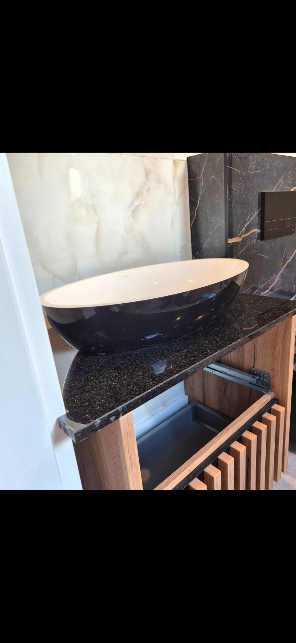 Nowoczesna łazienka: czarna, owalna umywalka nablatowa na blacie z ciemnego granitu, drewniana szafka z lamelami, marmurowe płytki na ścianie.