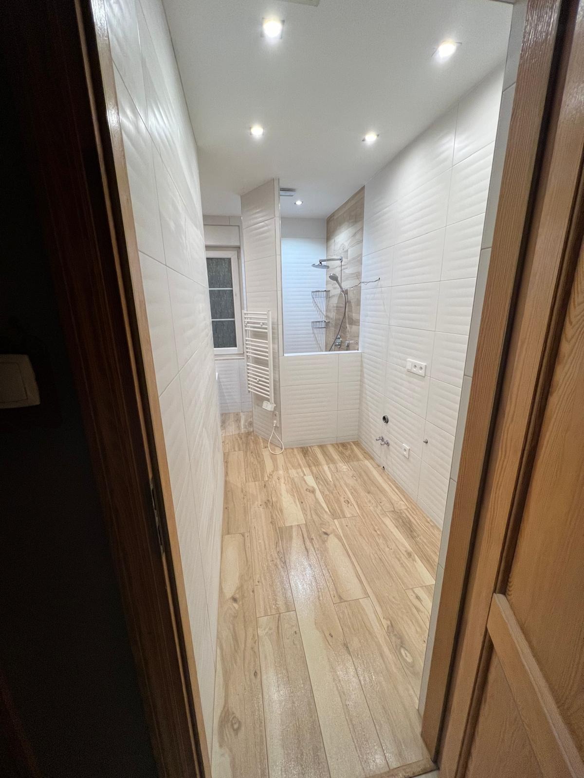 Wykończona łazienka z drewnopodobną podłogą i białymi płytkami ściennymi, widok zza uchylonych drzwi. Nowoczesny grzejnik i kabina prysznicowa w tle.