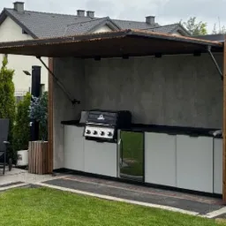 Zadaszona kuchnia letnia z grillem i szafkami, wykończona drewnem i betonem, z widocznym fragmentem ogrodu i domu w tle. Elementy drewniane pionowe.