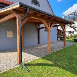 Drewniana pergola przy domu z szarym tynkiem, widoczny system odprowadzania wody. Konstrukcja oparta na czterech słupach, zadaszenie z desek, kostka brukowa na podłożu.